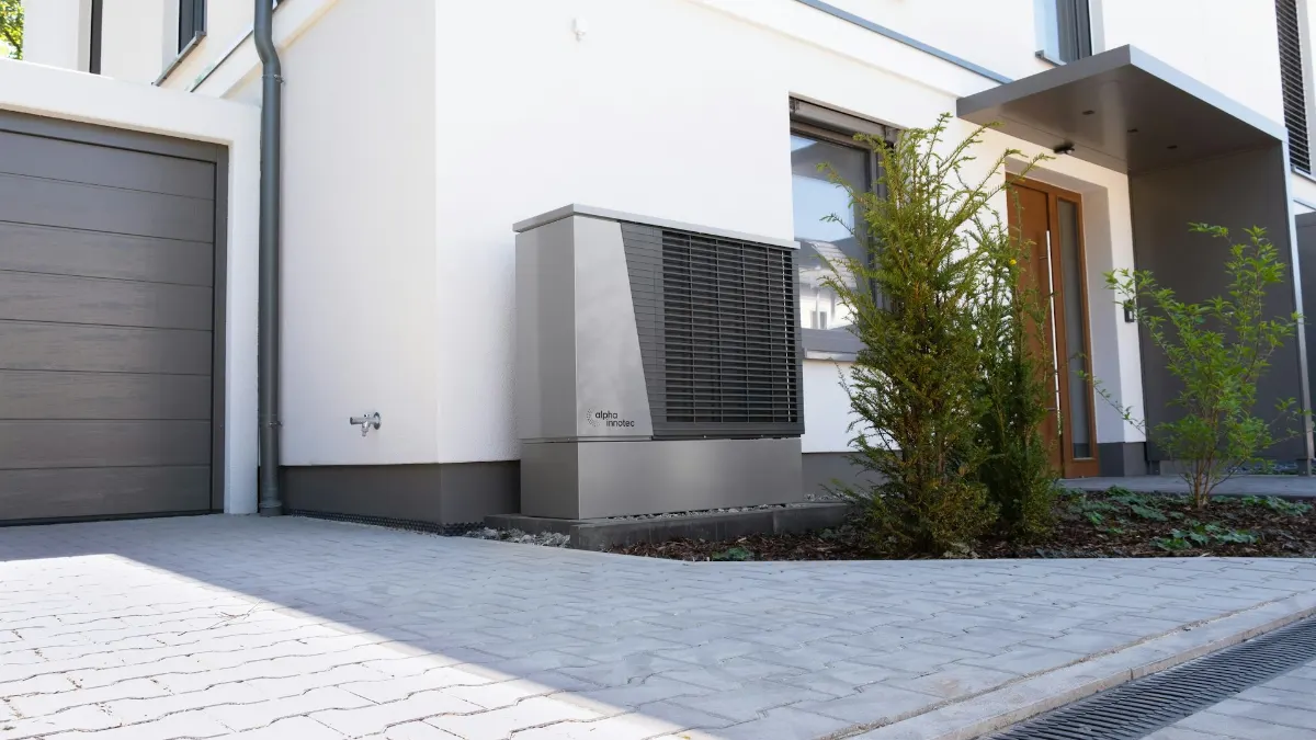 Unité extérieure de pompe à chaleur installée au pied d’une maison blanche, à côté de l’allée du garage